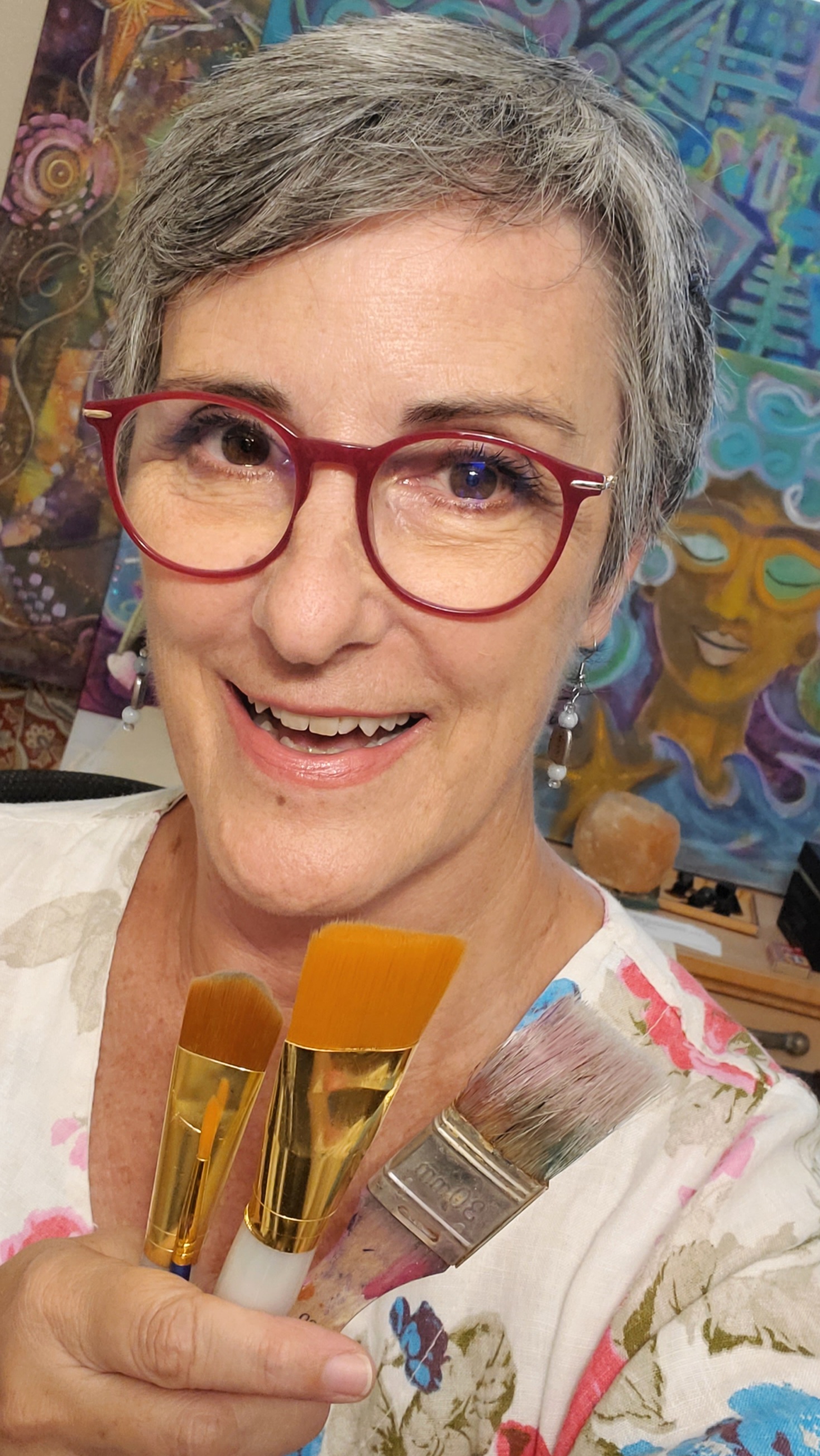 Nanette Saylor smiling in front of paintings and holding paint brushes